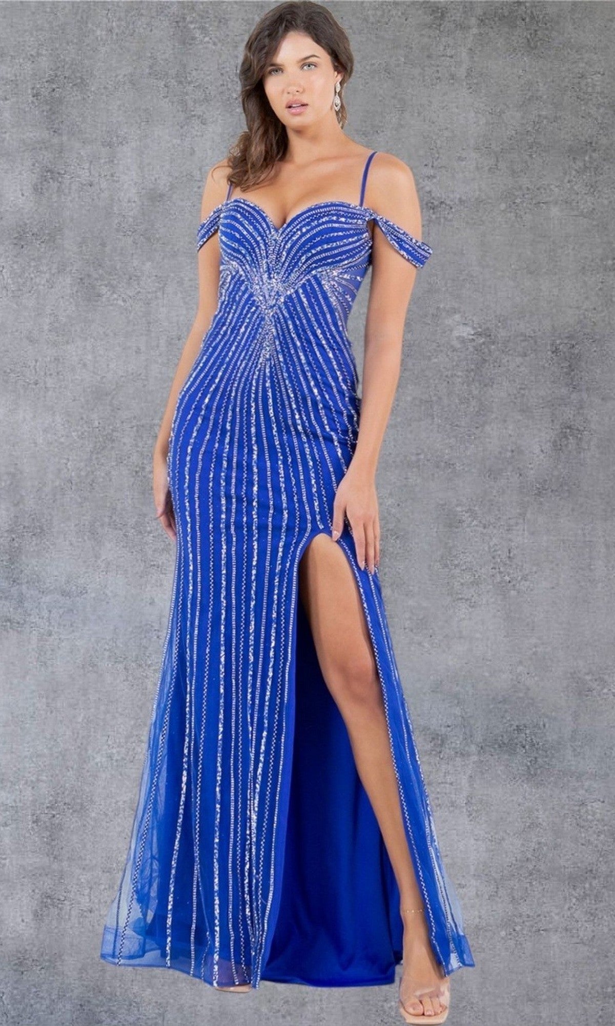 A woman wearing the Cinderella Couture 8269J long prom dress in royal, featuring sparkling blue beading, spaghetti straps, and a thigh-high slit, stands in front of a gray textured background.